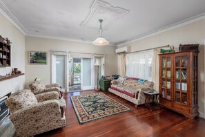 A spacious living room with floral-patterned armchairs, a wooden cabinet, and a large rug. - Albert Road Cottage: Comfy, vintage home near water & town  (East Bunbury)