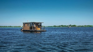 A houseboat with a small deck and a flag, floating on calm water. - Nice Houseboat in Kinrooi for Rent (Kinrooi)