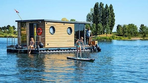 A floating houseboat with a wooden exterior, a small deck, and a lifebuoy. - Nice Houseboat in Kinrooi for Rent (Kinrooi)