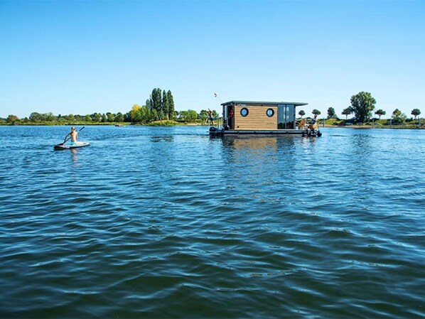 A houseboat with a wooden exterior and two circular windows, floating on calm water with a person paddleboarding nearby. - Nice Houseboat in Kinrooi for Rent (Kinrooi)
