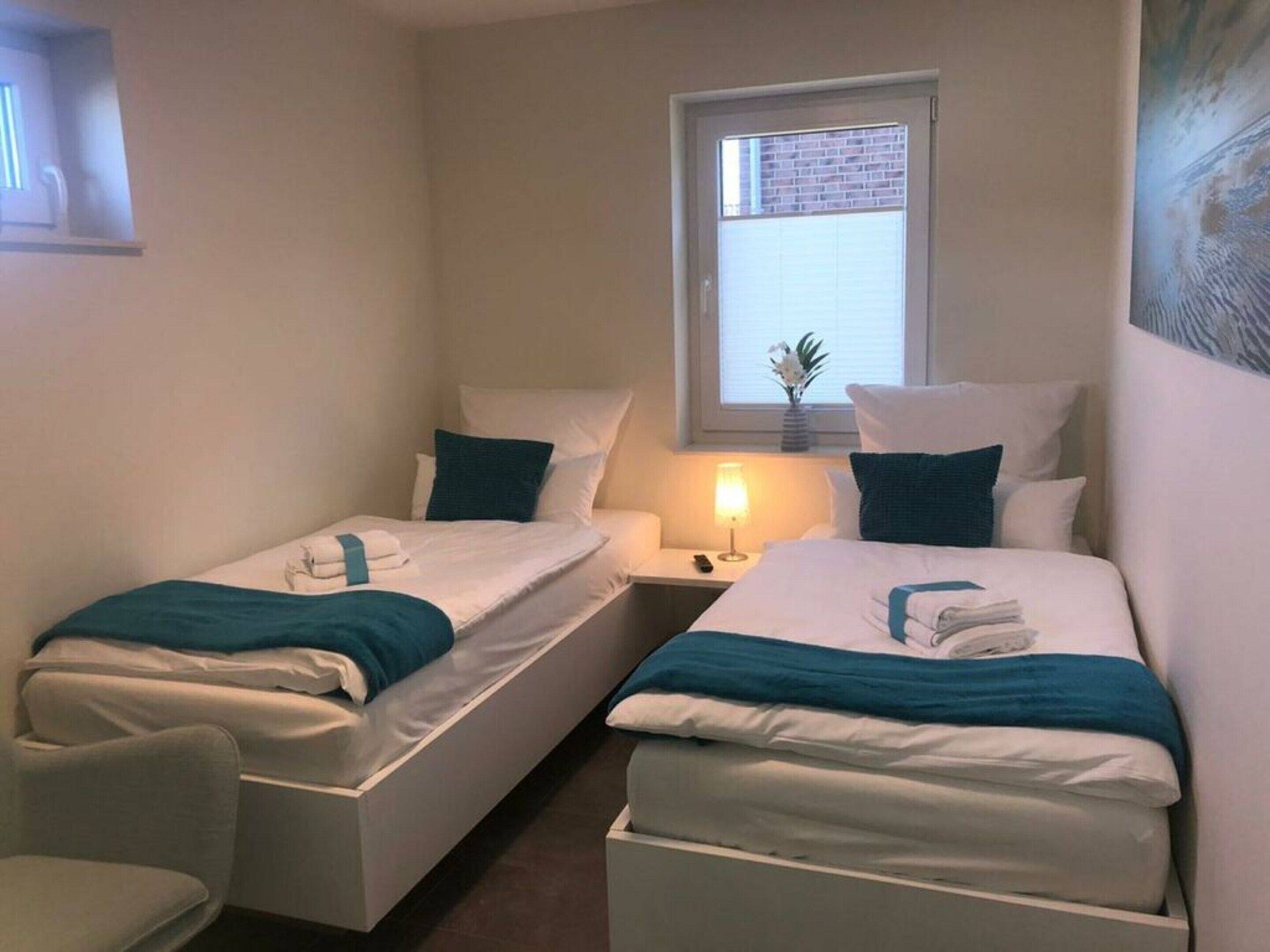 Une chambre double avec deux lits simples, chacun agrémenté d’une couverture et d’un oreiller de couleur turquoise, une table de chevet avec une lampe et une fenêtre donnant sur l’extérieur.