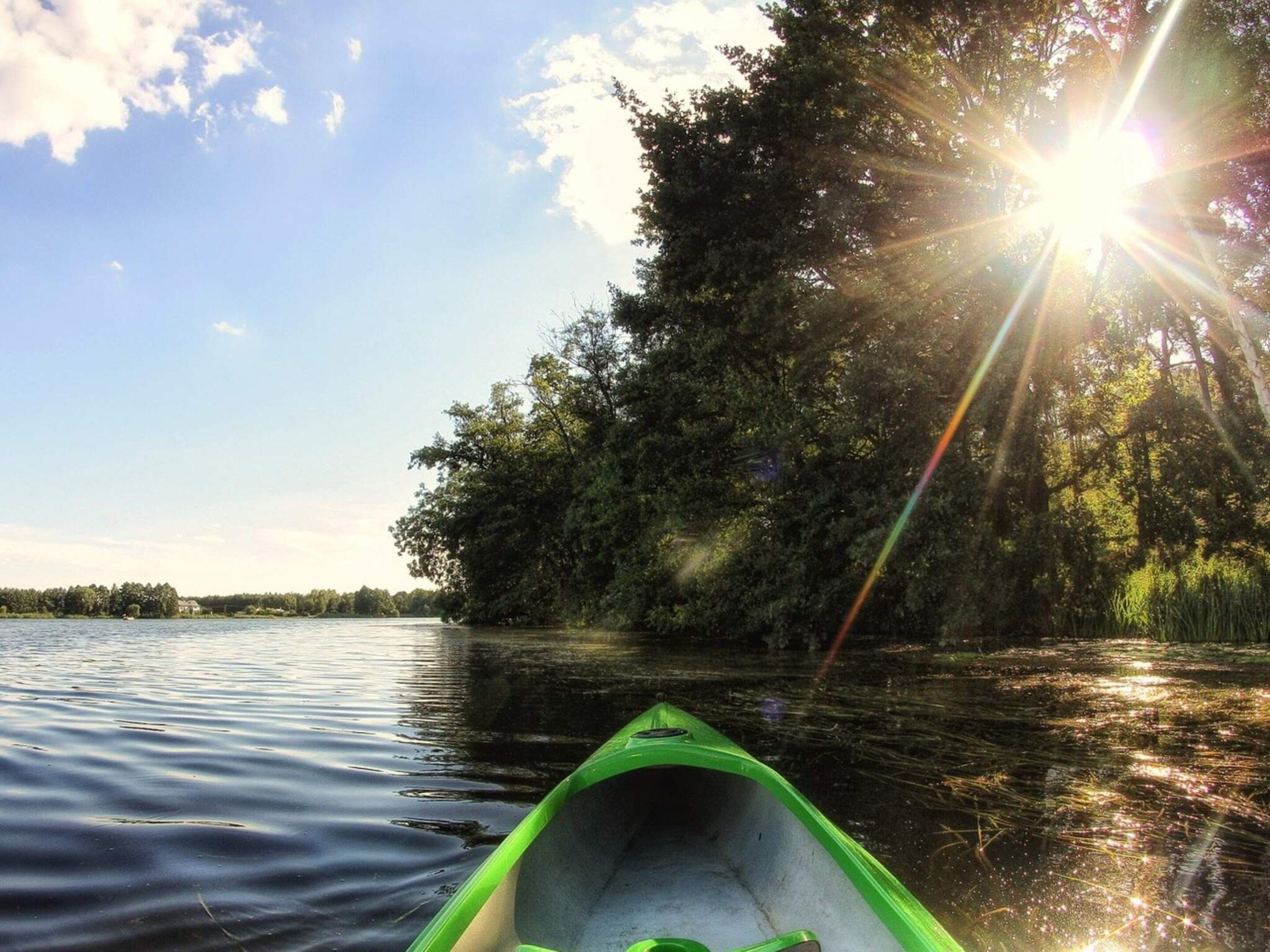 A green canoe on calm water with a forested shore and sunlight shining through the trees.