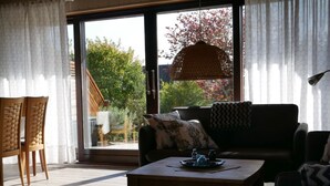 A cozy living room with a sofa, a coffee table, and a dining table. There is a large window with curtains, a wicker hanging light, and a view of a garden and trees. - Long Comfortable Holiday Residence (Cuxhaven)