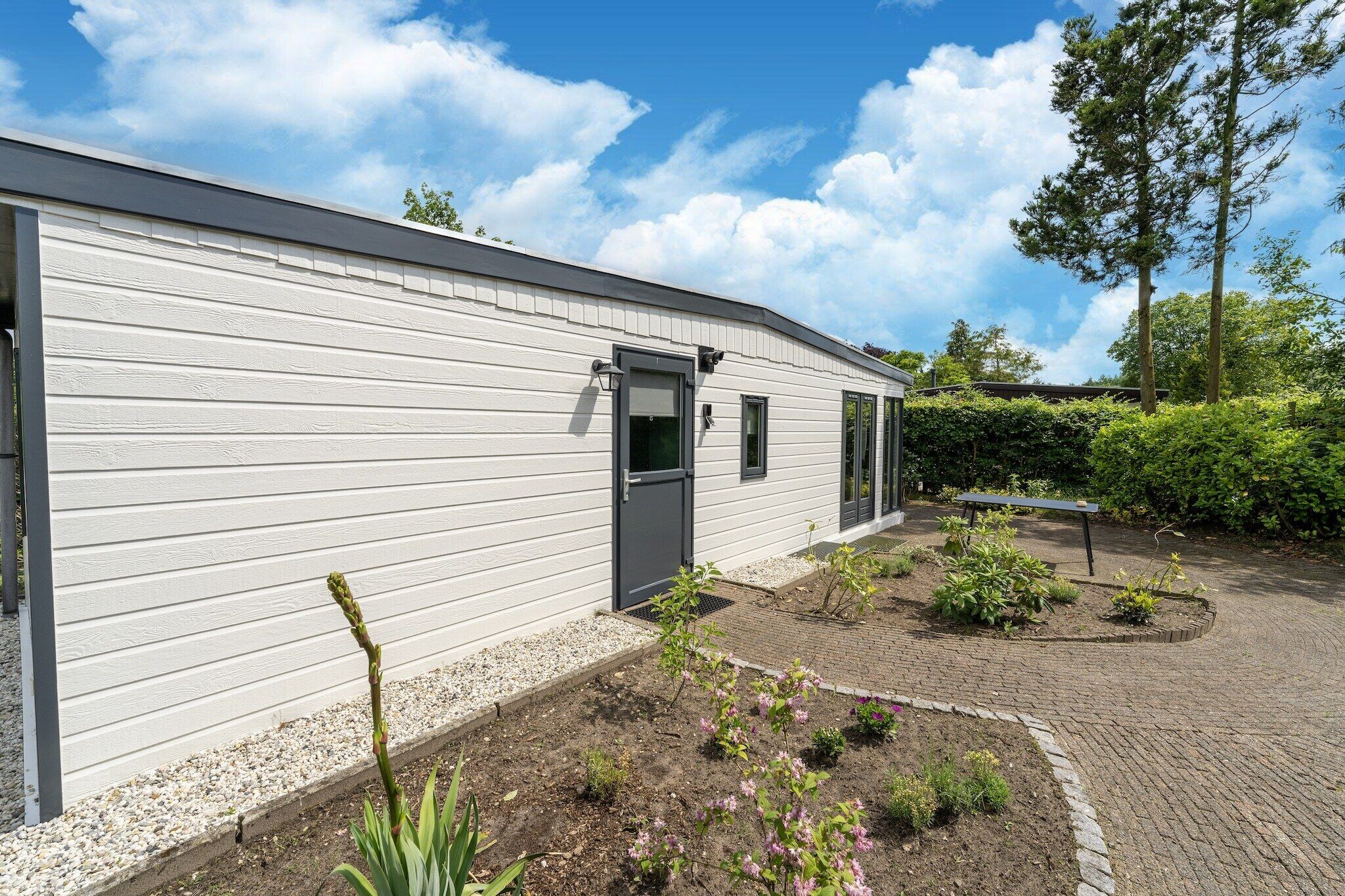 Ein weißes Gebäude mit einer grauen Tür, ein kleiner Garten mit Pflanzen und einer Bank, und ein klarer, blauer Himmel mit Wolken.