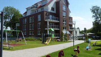 A playground with swings, a slide, and a climbing structure in front of a multi-story building with balconies.