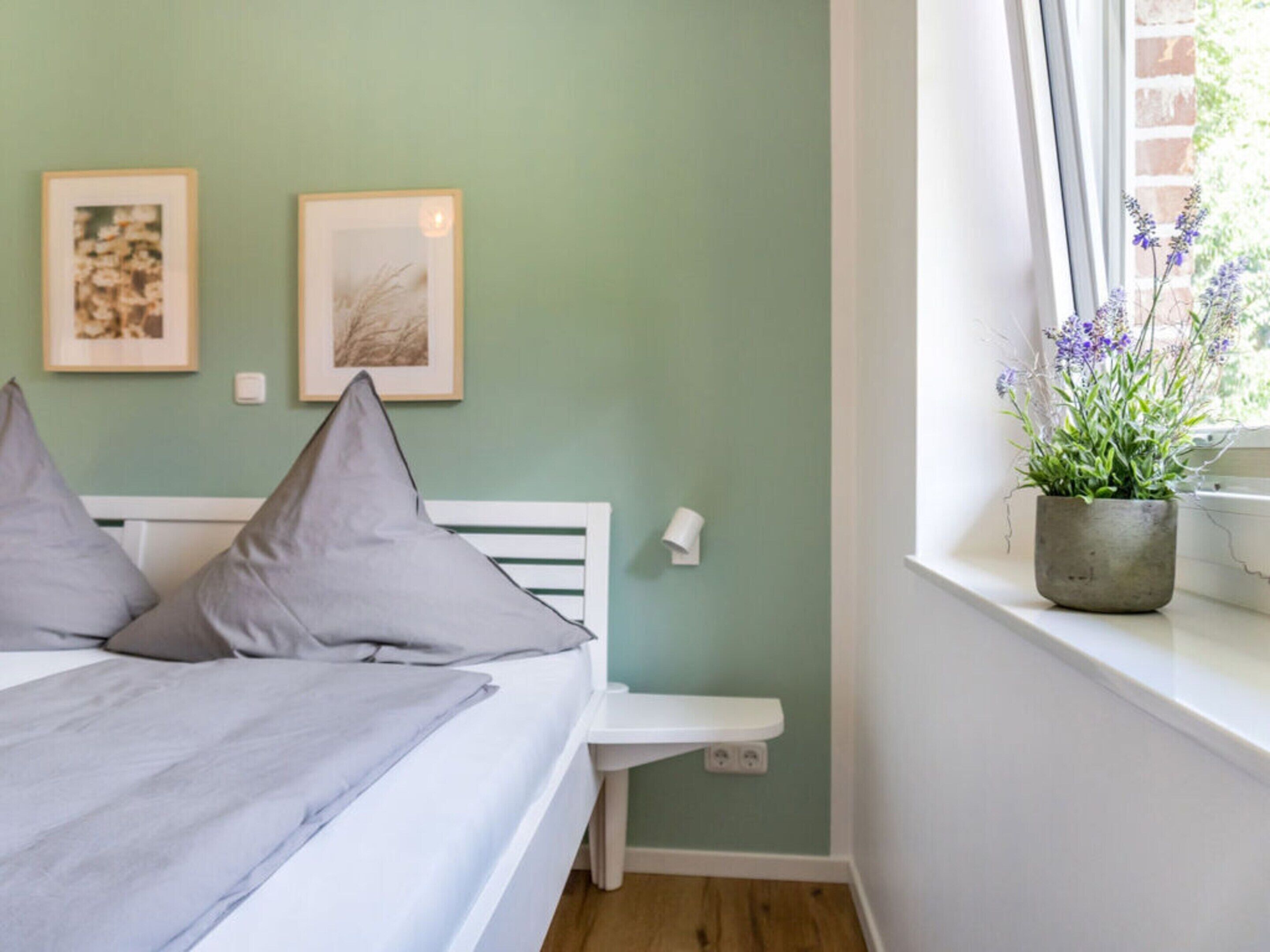 A bedroom with a bed, pillows, a bedside table, a window with a plant, and two framed pictures on the wall.