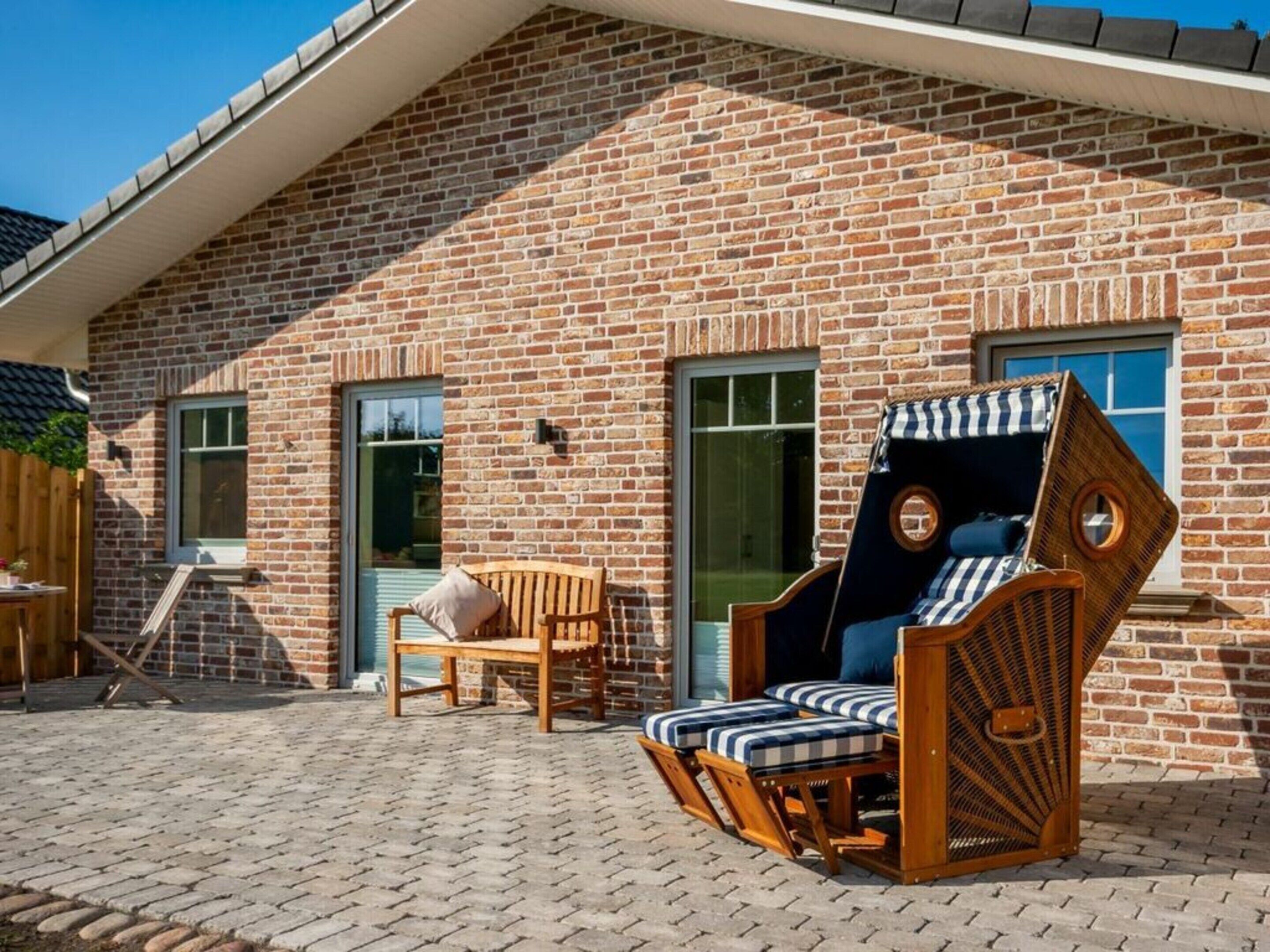 A brick house with a sloped roof, a wooden bench, and a wicker chair with striped cushions.