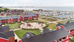 Un quartier résidentiel situé sur la côte, caractérisé par des bâtiments en briques rouges, un port de plaisance avec des bateaux et une aire de jeux centrale.