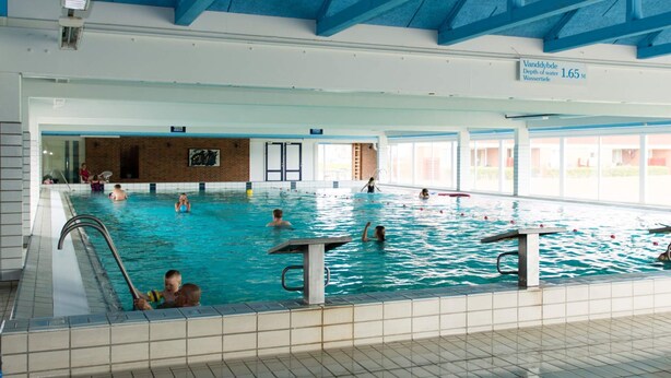 Une piscine intérieure dotée de plusieurs couloirs, d’un sol carrelé et d’un plafond haut avec des poutres bleues.