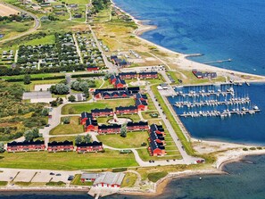 Luchtbeeld van een kustgebied met woonhuizen, meerdere gebouwen met rode daken, een jachthaven vol boten en een weg naar het strand.