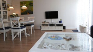 Una sala moderna con comedor, un televisor de pantalla plana montado en la pared y una mesa de centro de vidrio con objetos decorativos.