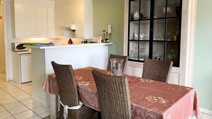 Een eethoek met een tafel voor vier personen, omringd door rieten stoelen, een kast met glazen deuren en een keuken op de achtergrond.
