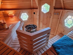 A wooden sauna room with hexagonal windows, a wooden bench, and a fire pit. - Strandperle Comfortable Holiday Residence (Kronsgaard)