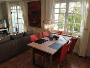 A modern living room with a sofa, a dining table with chairs, a TV, and a large window with a view of greenery. - Modern Apartment Near Seabeach (Binz)