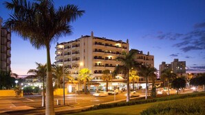 Un hotel de varios pisos con balcones, situado cerca de una calle con palmeras y farolas.