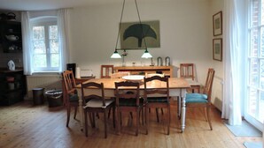 A dining area with a wooden table and chairs, a hanging pendant light, and a window with white curtains. - Old Zerwelin Forestry Company (Nordwestuckermark)