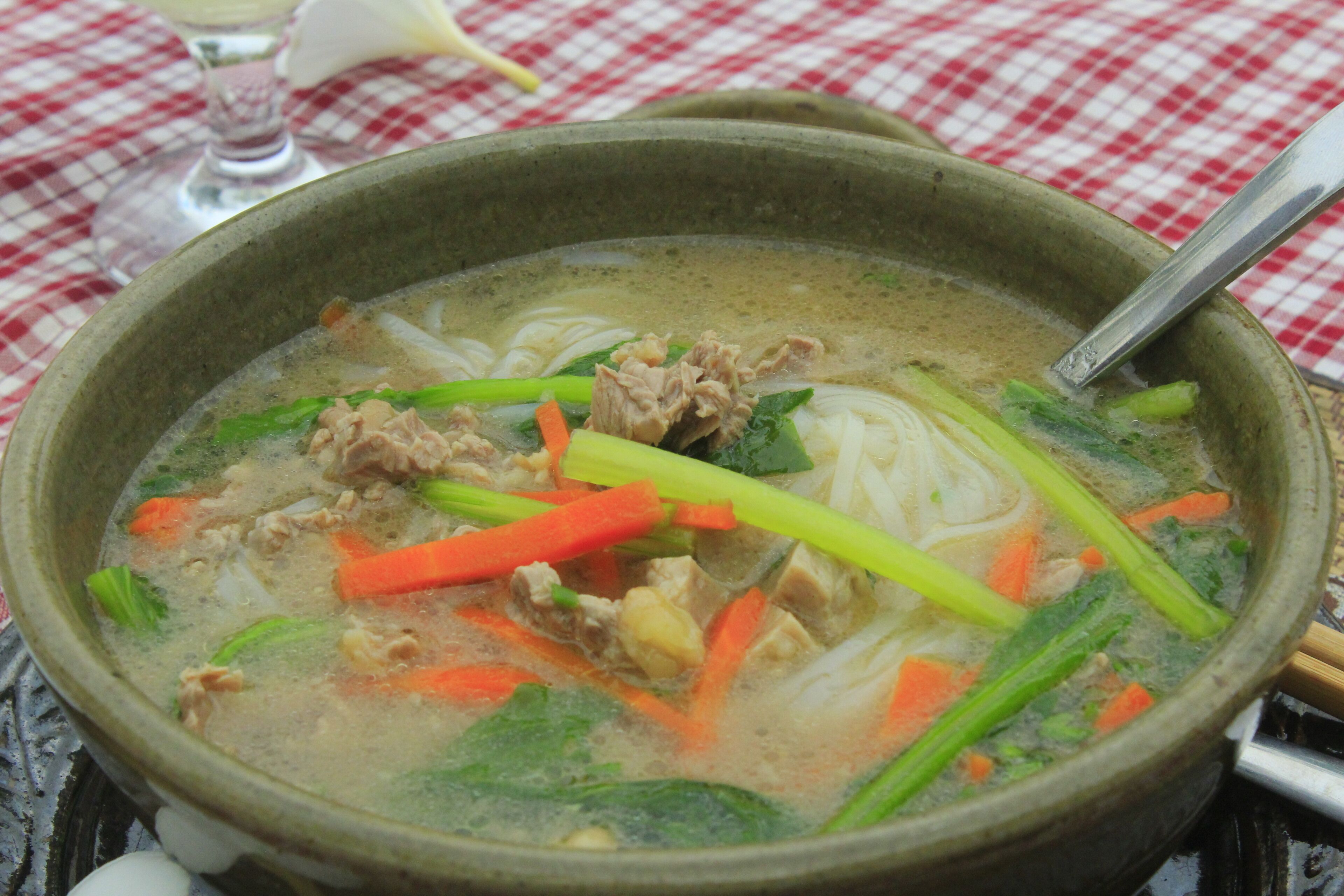 Un tazón de sopa con fideos, carne y verduras sobre una mesa con mantel a cuadros.