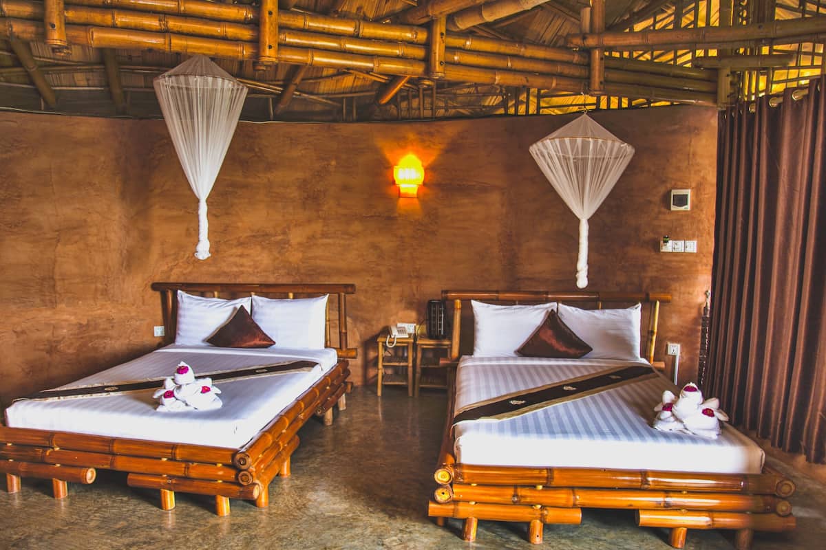 Two wooden beds with white linens and decorative pillows, a wall-mounted light, and a bamboo ceiling.