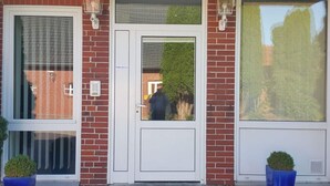 A red brick building with a white door and two windows on either side. There are two potted plants in front of the windows.