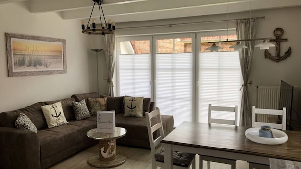 A cozy living room with a brown sofa, a small round table, and a wooden dining table. There is a painting on the wall and a window with curtains. - Dat Onnen's Hus - Burhave 16 (Butjadingen)