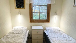 Une petite chambre à coucher composée d’une seule pièce, avec deux lits simples, une commode en bois et une fenêtre donnant sur un paysage verdoyant.
