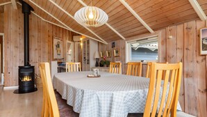 A cozy wooden interior with a central table, chairs, a fireplace, and a view of the sea.
