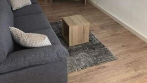 A living room with a grey sofa, a wooden coffee table, a television on a wooden stand, a wooden door, and a window with white curtains. - Ferienhof Schonmetzler (Altusried)