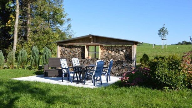 A stone building with a covered patio, outdoor seating, and a view of a green field. - Ferienhof Schonmetzler (Altusried)