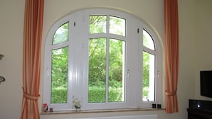 A living room with a large arched window, a radiator below, a television on a stand, and a sofa with a cushion. - Holiday Apartment Villa in the Park (Wangerooge)