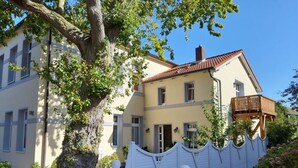 A two-story building with a light yellow facade, a red-tiled roof, and a white picket fence. - Holiday Apartment Villa in the Park (Wangerooge)