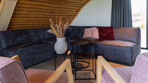 A modern loft-style room with a dark gray sectional sofa, a small round table, and a wooden chair. A vase with dried plants is on the sofa. - Peaceful Farmside Bungalow (Heeten)
