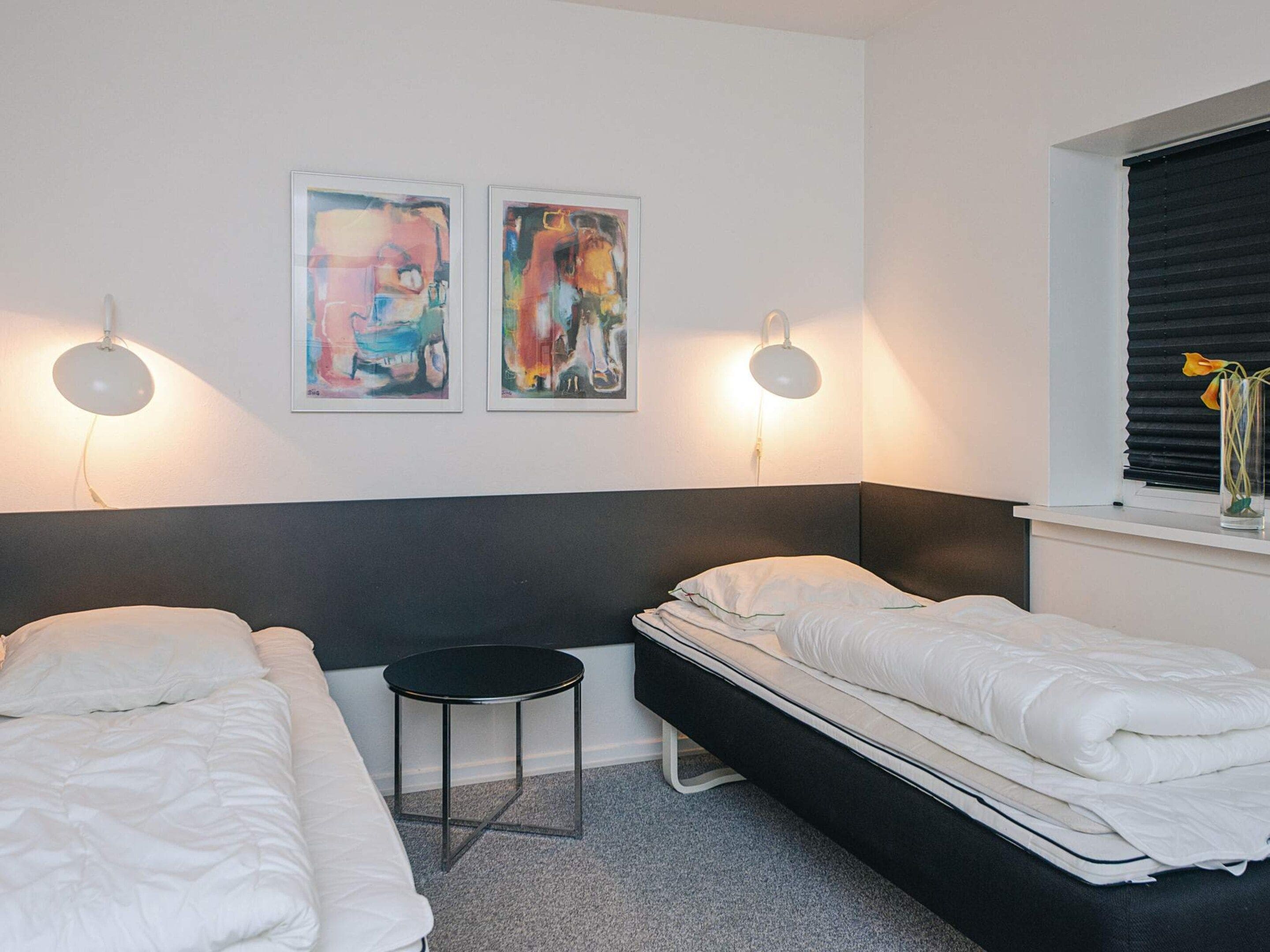 A hotel room with two beds, a small round table, two wall-mounted lamps, and a window with blinds.