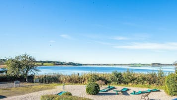 Vista sobre un lago con una zona de grava, sillones de jardín verdes y un cielo azul despejado.