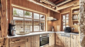 A rustic kitchen with wooden cabinets, a stone floor, and large windows overlooking a log storage area. - Felsenhutte Modern Retreat (Graen)