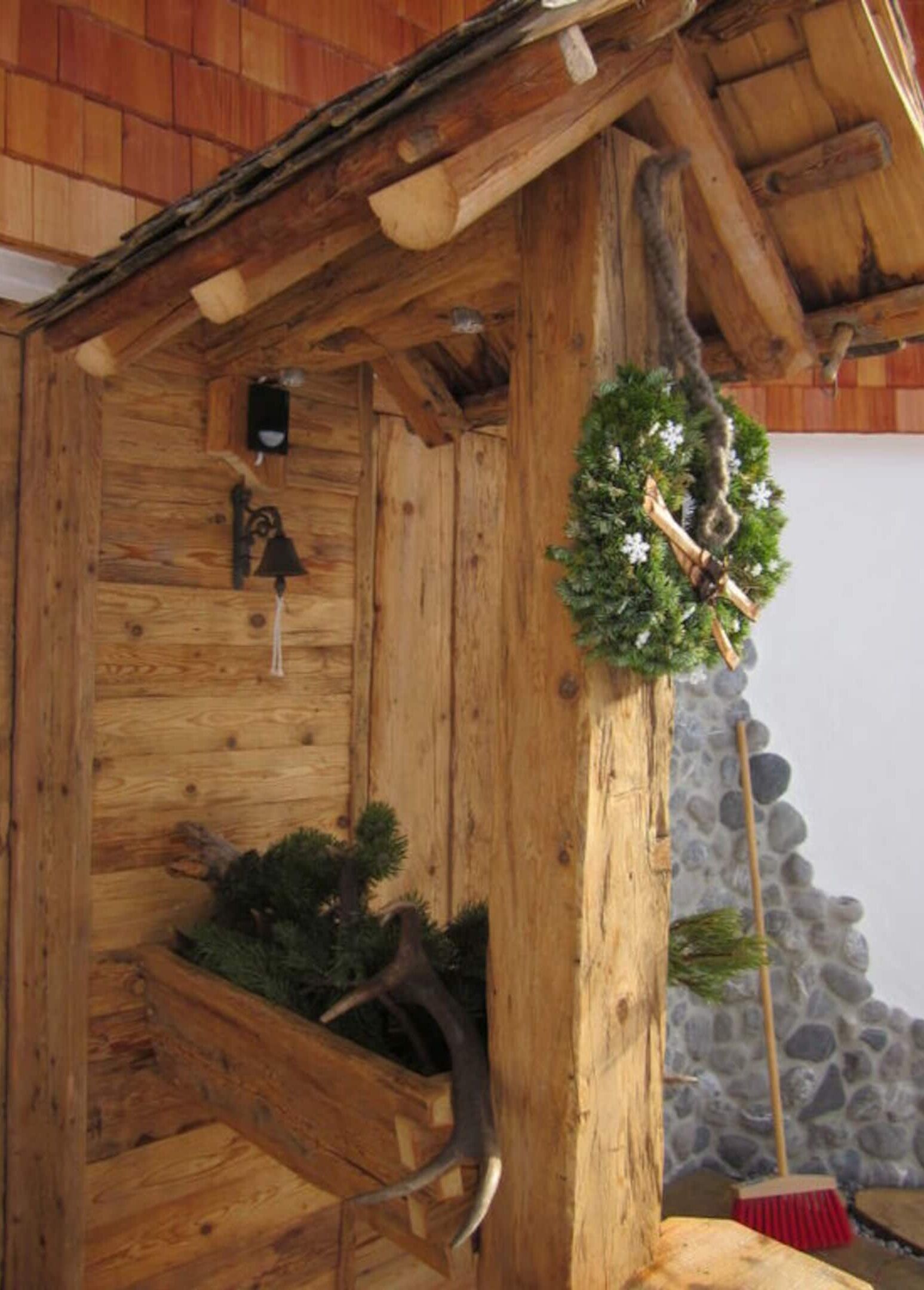 Eine Holzhütte mit einem Strohdach, eine Holzbank mit Grünpflanzen, einem Kranz und einer Steinmauer im Hintergrund.