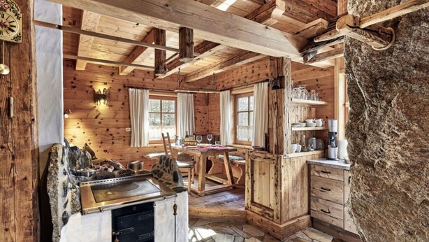 A rustic cabin interior with exposed wooden beams, a stone-tiled floor, a central dining table, and a stone fireplace. - Felsenhutte Modern Retreat (Graen)