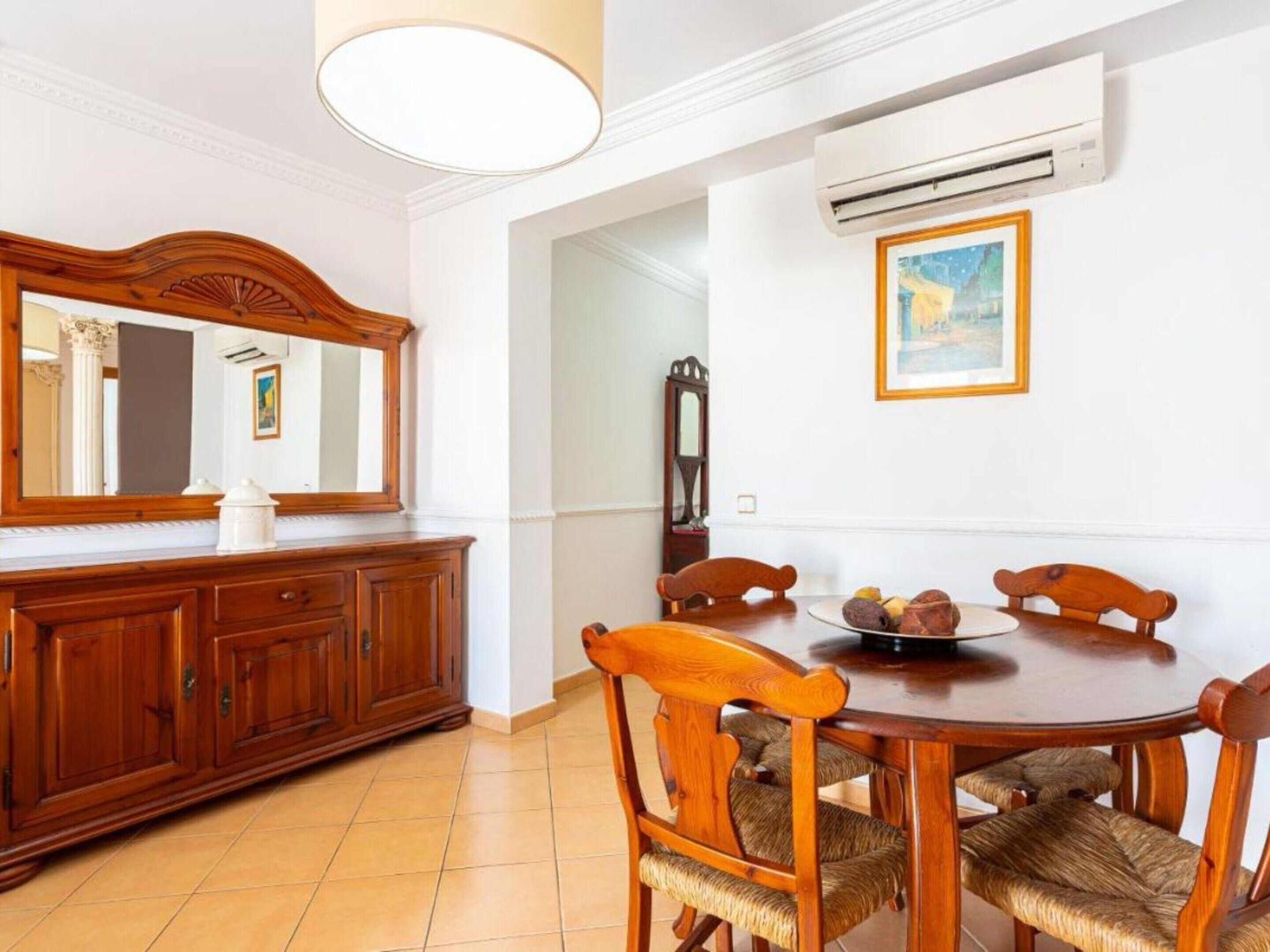 Un comedor con mesa y sillas de madera, un espejo, un aire acondicionado y un cuadro en la pared.