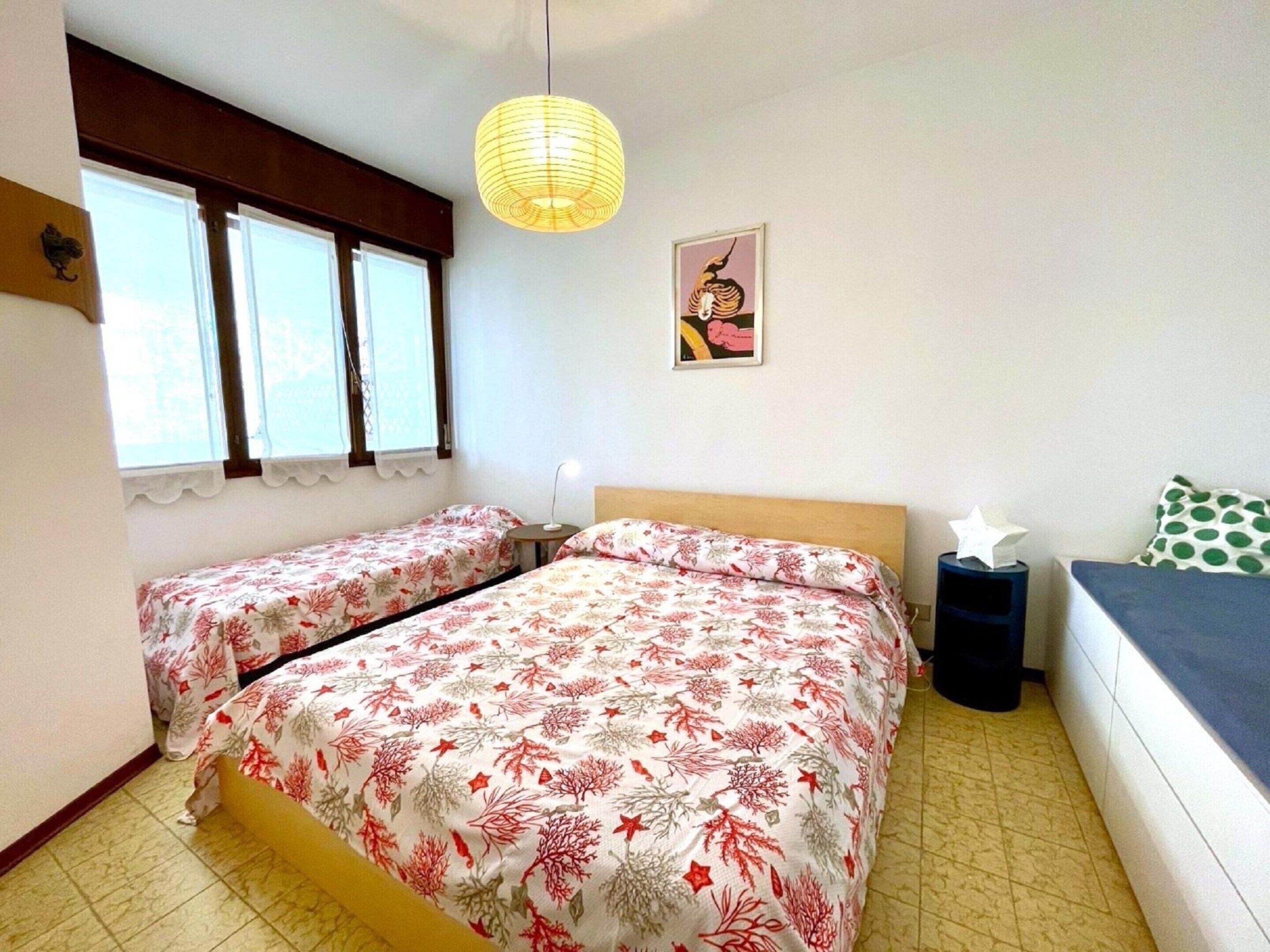 A bedroom with two single beds, a bedside table with a lamp, a window with curtains, a wall-mounted picture, and a hanging light fixture.