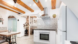 A modern kitchen with white cabinets, a built-in oven, and a stainless steel range hood. There is a wooden dining table with chairs and a bench. The kitchen has a tiled backsplash and wooden beams on the ceiling. - Stylishly Decorated Apartment With Private Terrace (Punat)