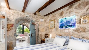 A bedroom with a stone wall, a bed with striped bedding, a nightstand, a window with a painting, and a view of the outdoors. - Stylishly Decorated Apartment With Private Terrace (Punat)
