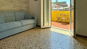 A room with a light blue sofa, a tiled floor, and a glass door leading to a balcony with a yellow wall and a table with chairs. - La Casa sul Mare (Vernazza)
