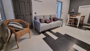 A living room with a grey sofa, a wicker chair, a dining table with chairs, and a patterned rug.