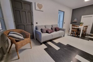 A living room with a grey sofa, a wicker chair, a dining table with chairs, and a patterned rug.