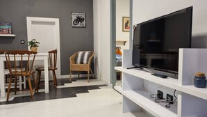 A living room with a TV, a dining table with chairs, a wicker chair, and a white door.
