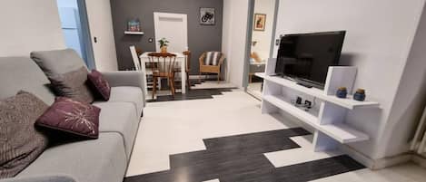 A modern living room with a grey sofa, a black and white patterned rug, a TV on a white stand, and a dining area with a table and chairs.