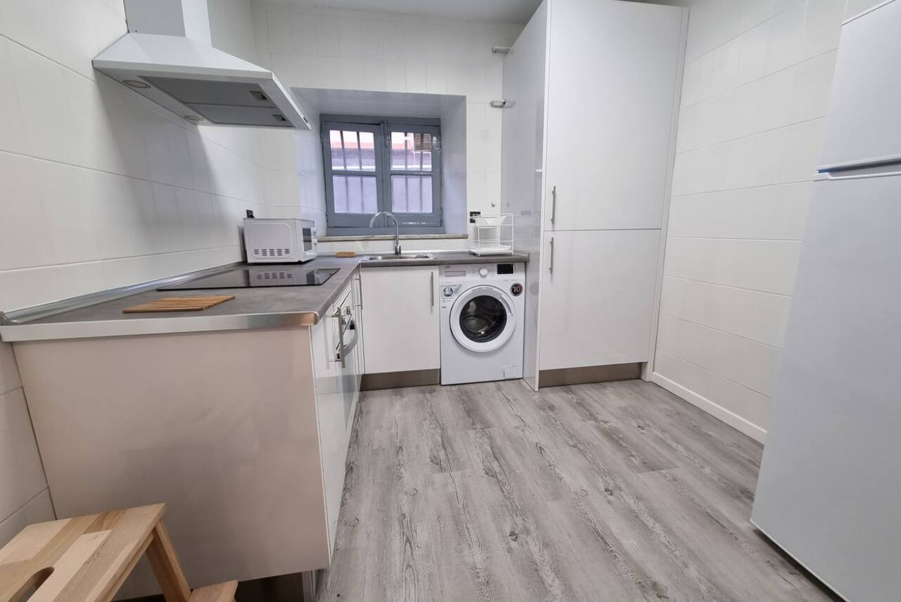 A compact kitchen with a washing machine, a window with a view, and a small wooden stool.
