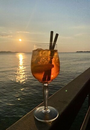 A glass of amber-colored beverage with a straw, placed on a balcony with a view of the sea and a sunset. - Blizina Mora II Modern Retreat (Vir)