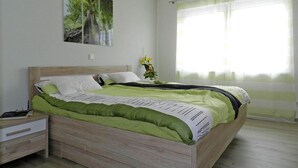 A bedroom with a wooden bed, a nightstand, a bedside table, a window with sheer curtains, and a painting on the wall. - Holiday Apartment Deck Boat (Butjadingen)