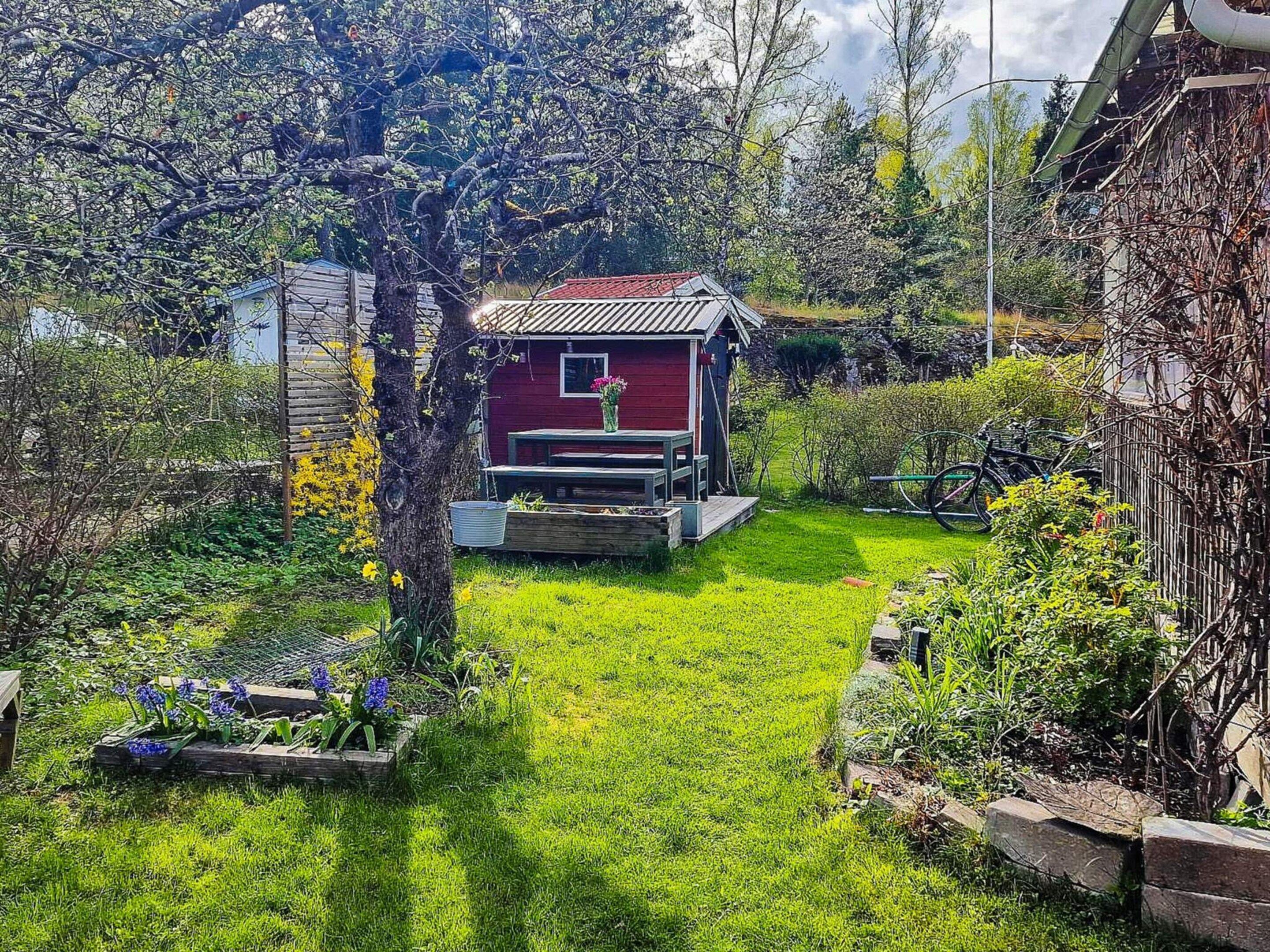 A red wooden house with a small porch, surrounded by a well-maintained garden with various plants and flowers.