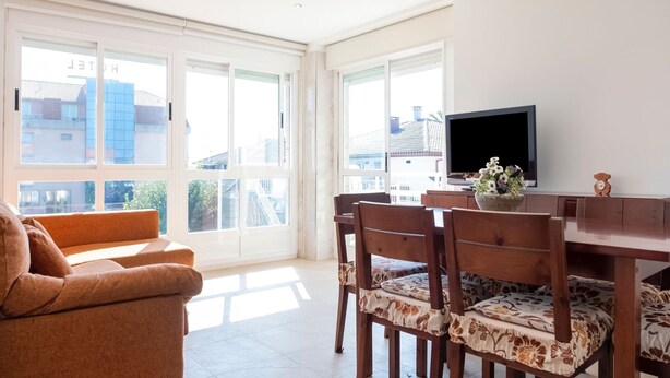 Ein helles Wohnzimmer mit einer Couch, einem Esstisch und Stühlen. Ein Fernseher steht auf dem Tisch und ein Fenster bietet Blick auf Gebäude.